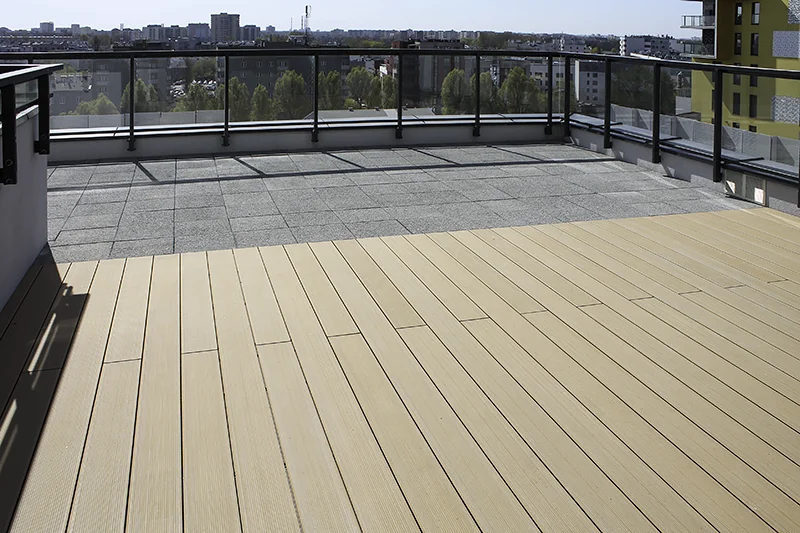 Jasne deski kompozytowe na tarasie dachowym z czarną balustradą, w tle panorama miasta