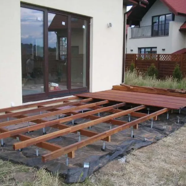 Ruszt z legarów na kotwach nad czarną agrowłókniną przy ścianie domu, z częściowo ułożonymi deskami tarasowymi
