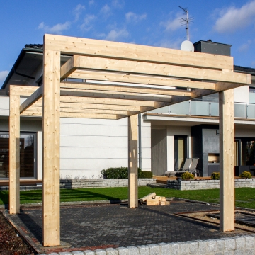 Pergola z jasnych belek klejonych przy nowoczesnym domu, ustawiona na kostce brukowej