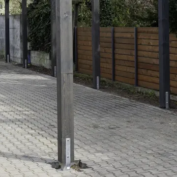 Rząd drewnianych słupów osadzonych na stalowych kotwach w kostce brukowej, w tle drewniane ogrodzenie