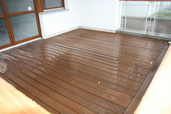 Balkon wyłożony ciemnobrązowymi deskami tuż po olejowaniu, powierzchnia jeszcze mokra, w tle drzwi tarasowe i balustrada