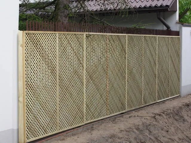 Wysokie kraty o gęstych oczkach ustawione w jednej linii przy białym murze, na nieurządzonym jeszcze terenie