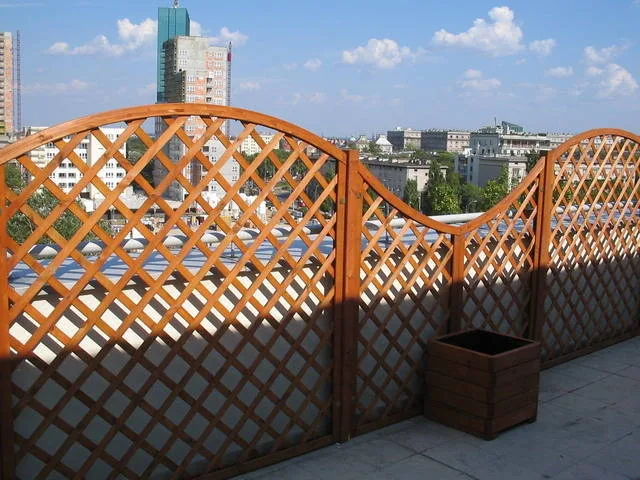 Kraty łukowe osłaniające taras na dachu, obok drewniana donica, w tle panorama miasta