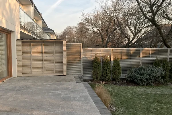 Ogrodzenie posesji z przęseł żaluzjowych ciągnące się wzdłuż trawnika, przy domu zabudowa z tego samego wzoru