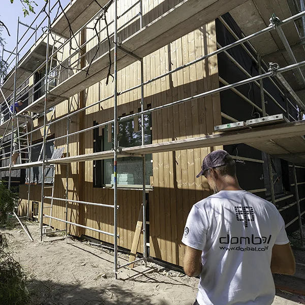 Monter w koszulce z logo Dombal przed elewacją z pionowych desek w trakcie montażu, wzdłuż ściany stoi rusztowanie