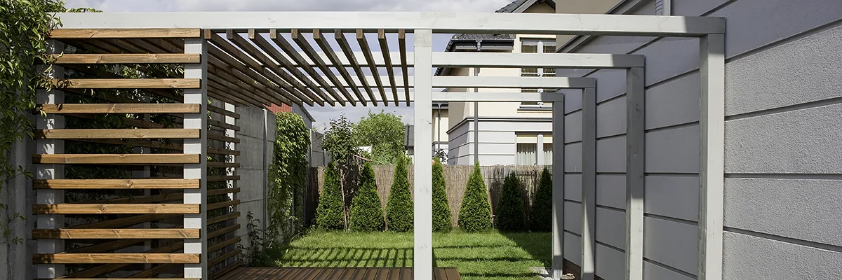 Pergola z poziomych listew przy jasnym budynku, listwy tworzą ażurową ścianę i dach, za nią rząd wysokich tuj