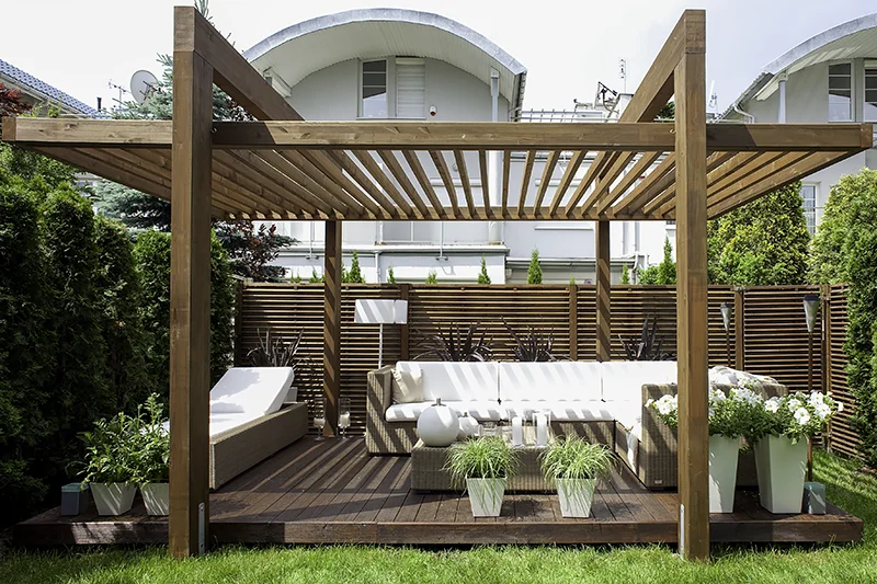 Pergola z ażurowym dachem z poprzeczek nad strefą wypoczynkową z leżankami, po bokach ekrany z poziomych desek, wokół donice z zielenią