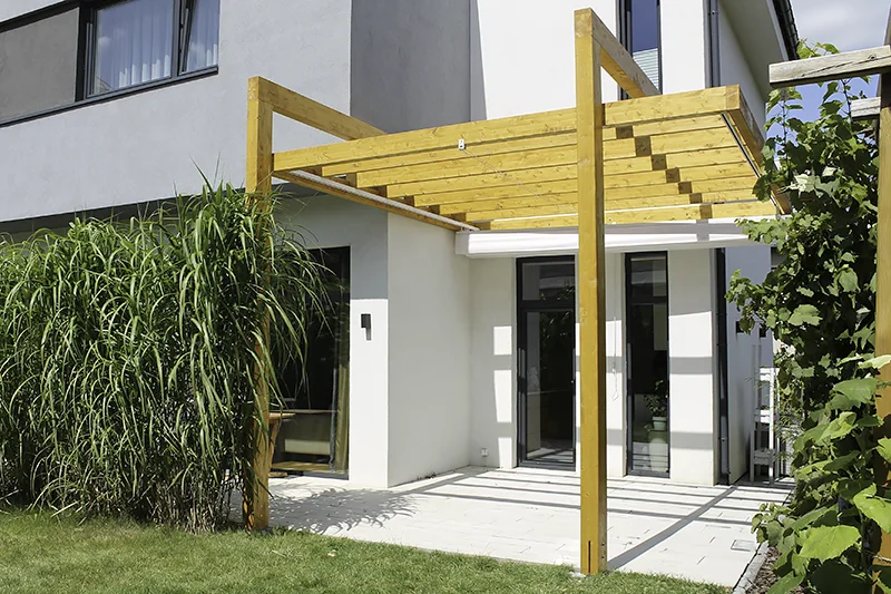 Pergola z jasnego drewna zamocowana do ściany budynku nad wejściem, obok wysokie trawy ozdobne i drewniana kratka na pnącza