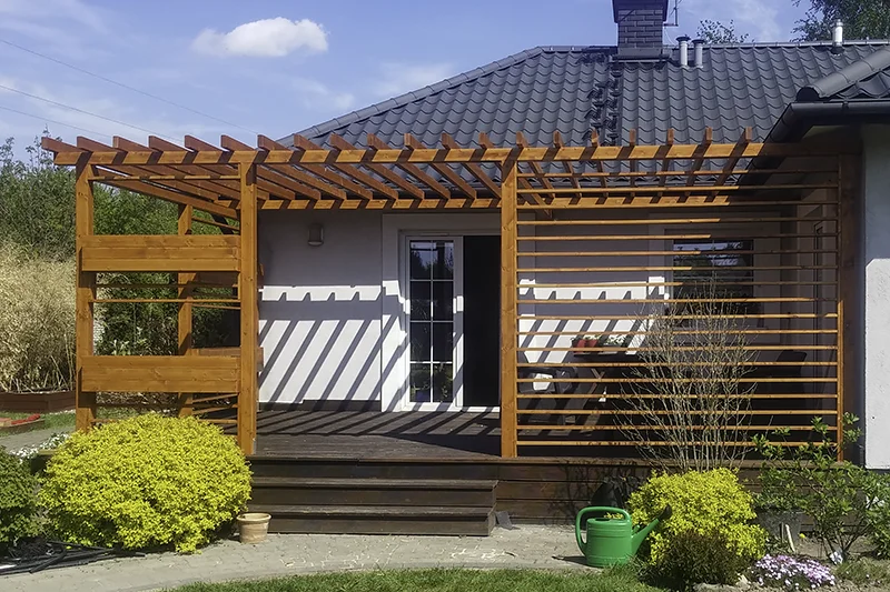 Pergola z jasnego drewna przy białym domu z ciemnym dachem, dach z gęsto ułożonych poprzeczek, przed nią przystrzyżone krzewy