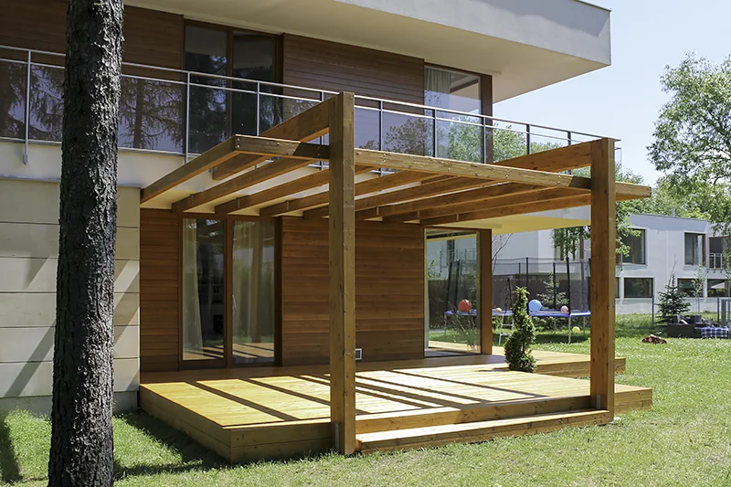 Pergola mocowana do ściany nowoczesnego domu ze szklaną balustradą, ażurowy dach z poprzeczek, pod nią drewniany podest i trawnik