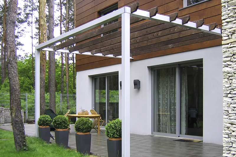 Pergola z ciemnego drewna przy domu z kamiennym cokołem, pod nią taras z płyt i donice ze strzyżonymi krzewami
