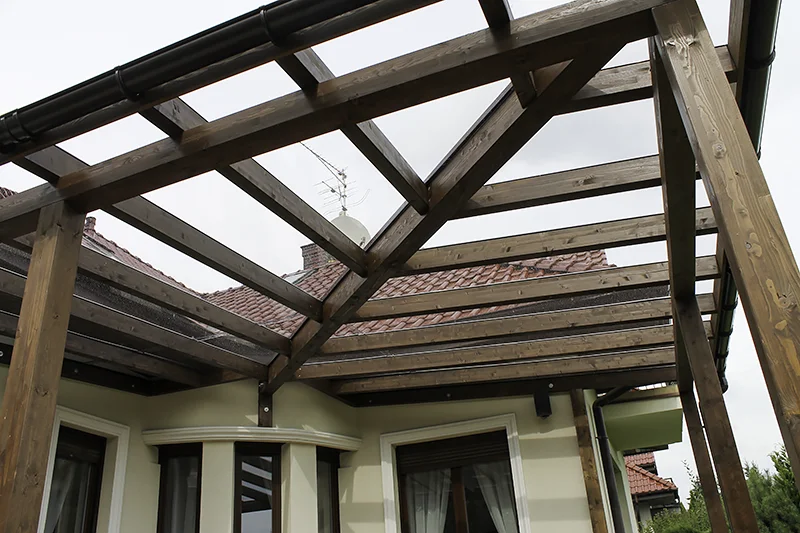Widok od spodu na konstrukcję zadaszenia: krokwie i płatwie z ciemnego drewna, między nimi przezroczyste pokrycie, w tle dach z dachówki